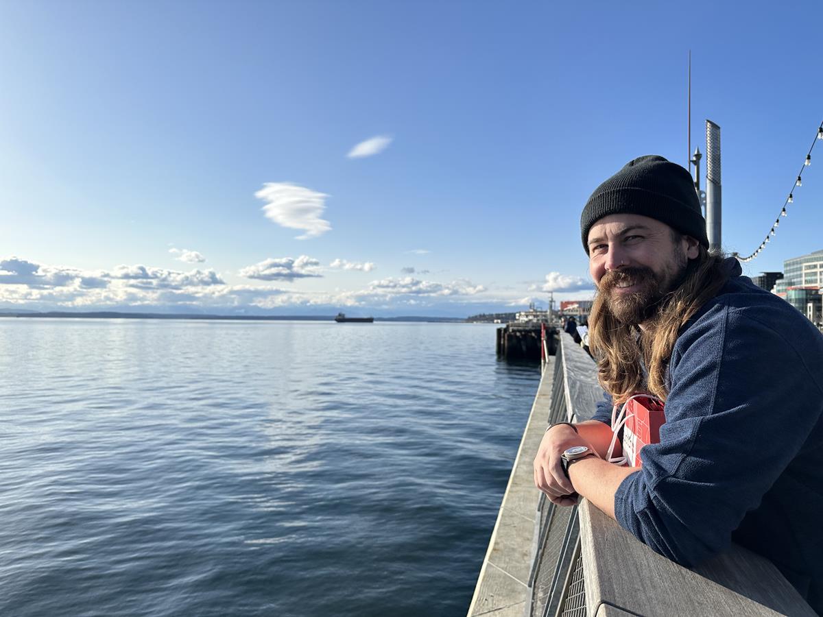 Right Foreground: A man with long hair and a beard smiles at the camera while leaning on a railing. Background: Blue skies and the waters of Elliot Bay, a ship can be seen far on the horizon.